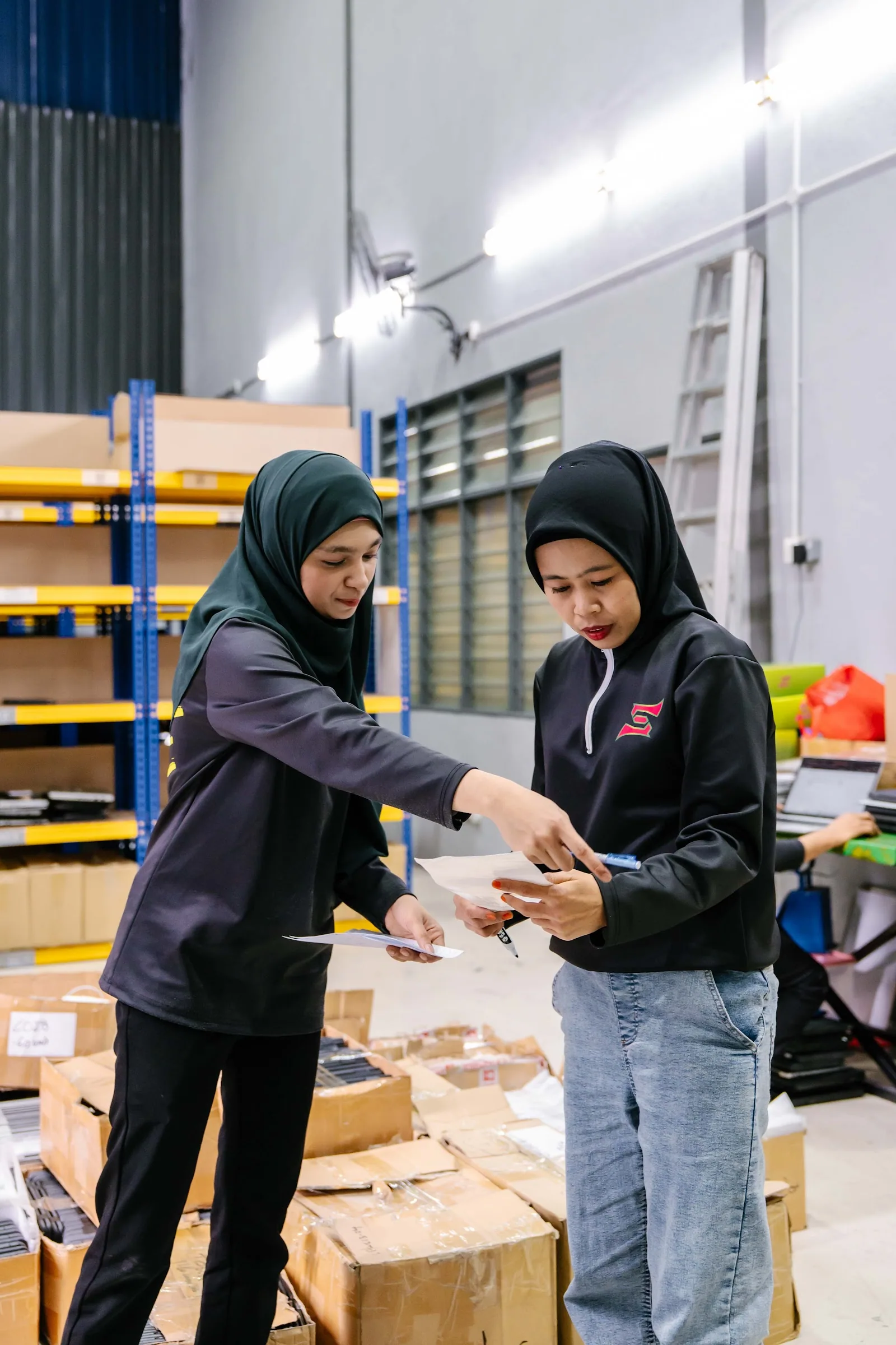 Two SCE staff checking refurbished laptop paperwork against racked stock in the Cheras warehouse
